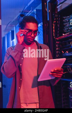 young african american programmer adjusting eyeglasses while looking at digital tablet in data center Stock Photo