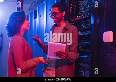 smiling african ameican engineer with digital tablet gesturing while talking with colleague near server Stock Photo