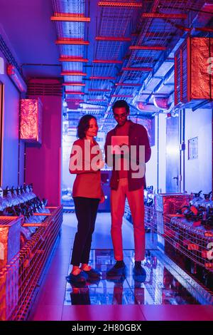 interracial technicians looking at digital camera while standing in technical equipment room in neon light Stock Photo