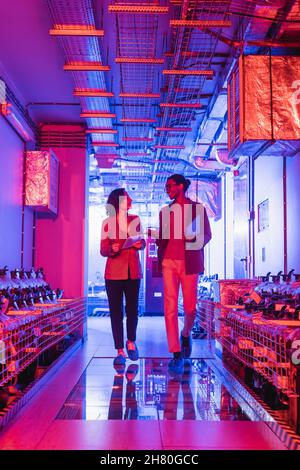 multiethnic engineers with gadgets talking while walking in technical equipment room in data center Stock Photo
