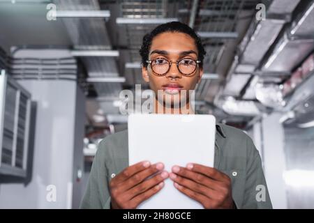young african american programmer holding digital tablet while looking at camera in data center Stock Photo