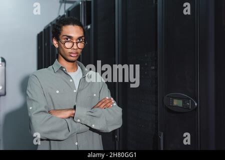 african american programmer in eyeglasses standing with crossed arms and looking at camera in data center Stock Photo