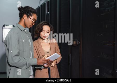 young programmer pointing at digital tablet while working with african american colleague in data center Stock Photo