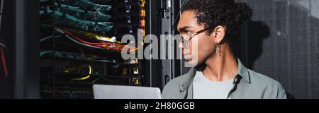 african american engineer with laptop looking at wires in server while working in data center, banner Stock Photo