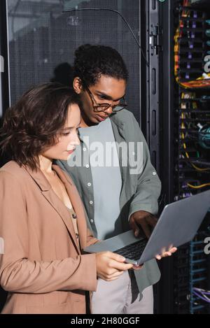 couple of multiethnic engineers looking at laptop while checking server in data center Stock Photo