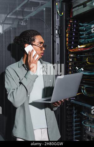 african american technician with laptop talking on smartphone while checking server in data server Stock Photo