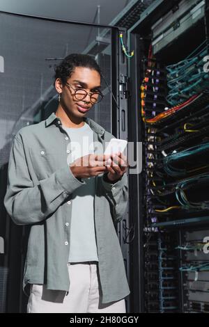 smiling african american programmer using mobile phone while making diagnostics of server in data center Stock Photo