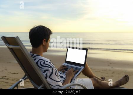 Lifestyle of young Asian man blogger working and using keyboard on laptop while sitting on the beautiful beach, freelance travel on holiday summer dur Stock Photo