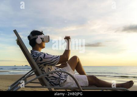 Young Asian man using virtual reality glasses for business meeting on the tropical beach over beautiful sea and sky background Stock Photo
