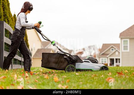 a young caucasian woman wearing overalls and noise blockers is operating a lawnmover on grass in late autumn. There are fallen leaves and a suburban n Stock Photo