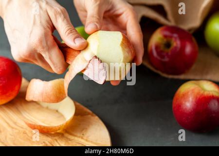 apples of different varieties are seen on dark stone and wooden background.  A woman is peeling the skin of an apple using a fruit knife. Healthy eati Stock Photo
