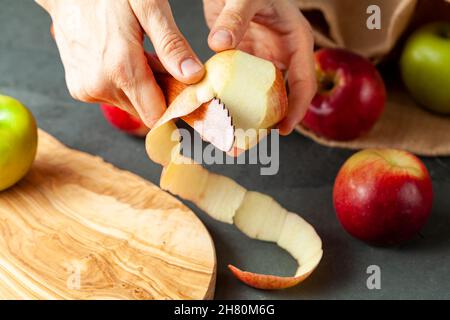 apples of different varieties are seen on dark stone and wooden background.  A woman is peeling the skin of an apple using a fruit knife. Healthy eati Stock Photo
