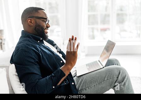 Glad to see you. Pleased young bearded African-American guy in casual wear waving hand into webcam, greeting participants of virtual meeting, using laptop for video connection on distance, side view Stock Photo