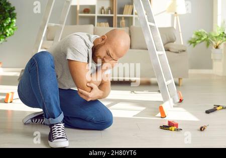 Man who just fell off ladder at home is sitting on floor and touching his hurt elbow Stock Photo