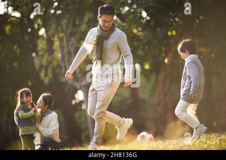 asian father and son playing soccer outdoors in park while mother and daughter watching from behind Stock Photo