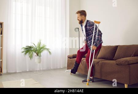 Man with leg injury leans on crutch and has difficulty getting up from couch at home. Stock Photo