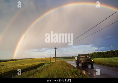 Old Russian truck on road in green fields with beautiful double rainbow and electric poles Stock Photo