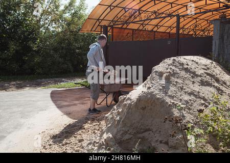 A 30-year-old caucasian man pours sand from a large pile of sand onto a garden wheelbarrow Stock Photo