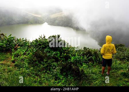 Boy in yellow rain jacket standing among plants, looking to crater lake Lagoa Funda das Lajes in fog, Flores Island, Azores, Portugal Stock Photo