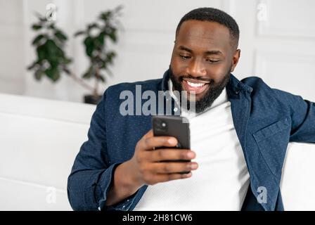 Happy African-American man is looking on the phone and smiling, in casual blue lying on the white sofa, black guy typing message, watching film, video Stock Photo