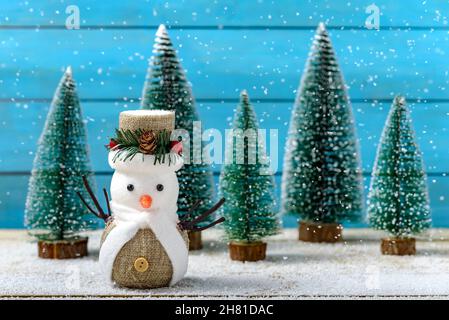 Christmas decoration snowman standing next to fir trees while it is snowing on a wooden table. Concept Christmas greeting card. Stock Photo