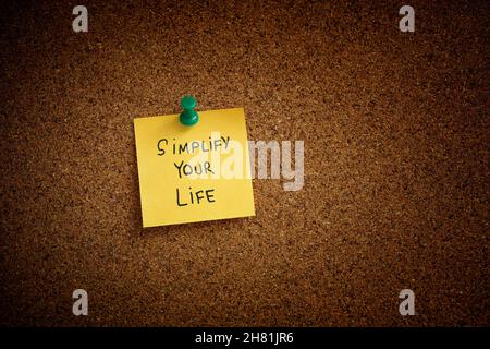 A yellow sticky note with the phrase Simplify Your Life written on it pinned to a cork board. Close up. Stock Photo