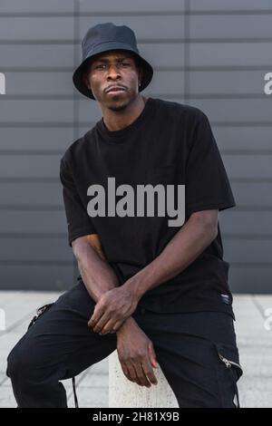 Vertical shot of a young black man in street-style clothes sitting on a pillar outdoors Stock Photo