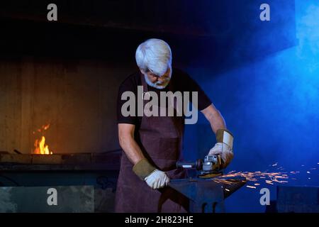 Experienced mature man in safety apron and gloves using industrial tool for processing steel at forge. Caucasian blacksmith creating some metal product at workplace. Stock Photo