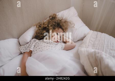 Cute charming curly-haired baby girl lying in bed on a white pillow. The concept of childhood. Stock Photo