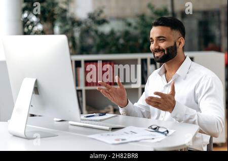 Friendly confident successful Indian man, businessman or mentor, sitting at his desk, wearing a white shirt, using computer for distant communication, talking to a colleague via video call, smiling Stock Photo