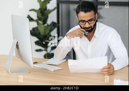Concentrated serious bearded Indian man, business man, in a formal attire, sitting at a table in the office, studying documents, analyzing a report and financial charts Stock Photo