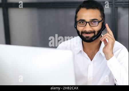 Head shot of a successful confident Indian man, operator of call center or business mentor. Businessman with glasses, uses headphones for online conference, looks at camera and friendly smiling Stock Photo