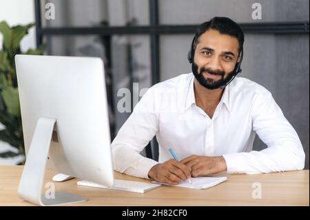 Online consultation, distance learning, online webinar. Indian positive man with glasses, takes notes during an online lesson or consultation, sits at a desk, formal dressed, smiles, looks at camera Stock Photo