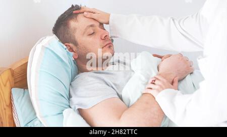Doctor  checking fever temperature of senior man lying on bed. Man resting at home feeling symptoms of the flu while wife checking fever by touching f Stock Photo