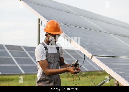 Professional technician in uniform using multimeter while checking voltage in solar panels. African american controlling production of green energy on station. Stock Photo
