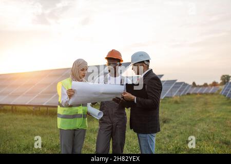 Group of three multiracial people in safety helmets having working meeting among solar station. Technician with engineers standing outdoors with blueprints in hands. Stock Photo