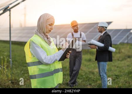 Side view of muslim woman in hijab writing on clipboard while standing among rows of solar panels. Two multicultural men in helmets standing behind with project plan in hands. Stock Photo