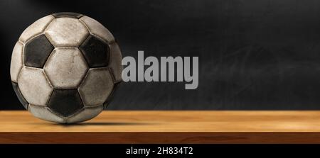 Closeup of an old leather soccer ball on a wooden table with empty blackboard on the background with copy space. Stock Photo