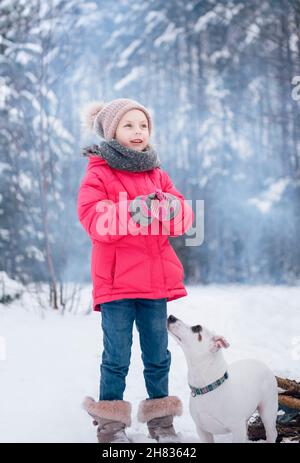 Little girl in a bright jacket plays in the winter snowy forest with her dog jack russell terrier Stock Photo