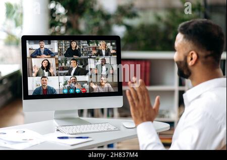 Video call, online conference. Over shoulder view of Indian man, on computer screen, talks with multinational group of successful business people, virtual business meeting, telecommunication concept Stock Photo