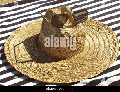 Beach hat and sunglasses on a white beach towel with black stripes on a sunny beach. Stock Photo