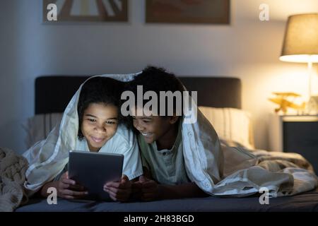 Positive black siblings lying under duvet on bed and using tablet while browsing internet at night Stock Photo