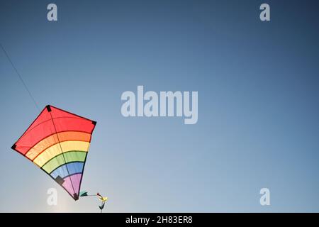 Multicolored striped kite flies in blue sky. Windy weather is great time to play with kite. Empty minimalistic background. Stock Photo