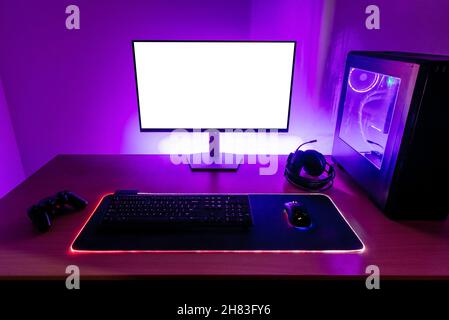 Desk with gaming setup. Display with isolated screen for mockup. Gaming PC, headset, keyboard, mouse and joypad on desk. Purple led light on wall Stock Photo