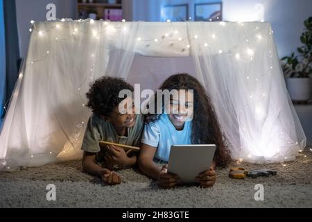 Cheerful African American brother and sister lying on carpet behind table covered with fabric and lights and using tablet Stock Photo