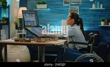 Disabled architect analyzing building model on touch screen monitor for construction layout. Engineer with handicap working on blueprint plan while sitting in wheelchair. Architectural equipment Stock Photo