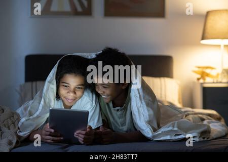 Positive black siblings lying under duvet on bed and using tablet while browsing internet at night Stock Photo