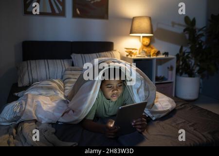 Concentrated Black boy lying under duvet and using tablet in dark nursery room Stock Photo