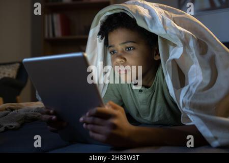 Concentrated African American boy with duvet on head lying in bed and using tablet while reading online book in darkness Stock Photo