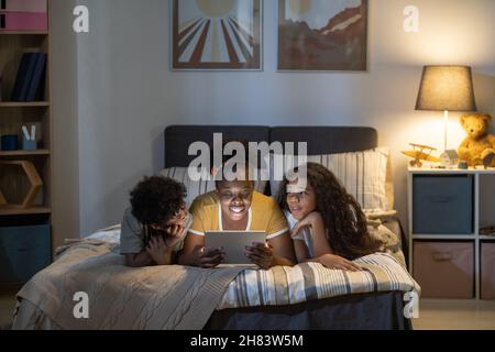 Happy curly-haired African American mother lying on bed and using tablet while reading fairy tale to kids before bedtime Stock Photo